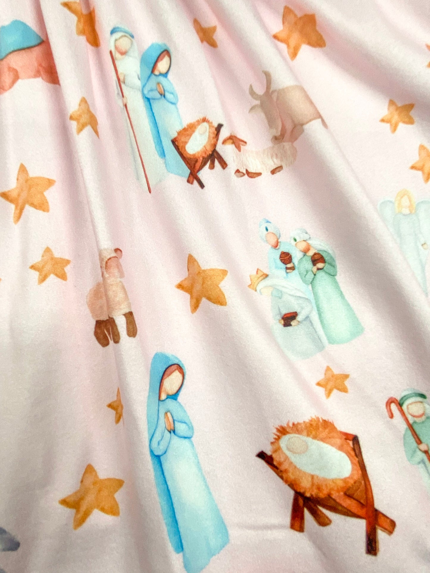 Detailed view of the pastel nativity scene print on a pink dress, featuring Mary, Joseph, baby Jesus in the manger, shepherds, animals, and gold stars.