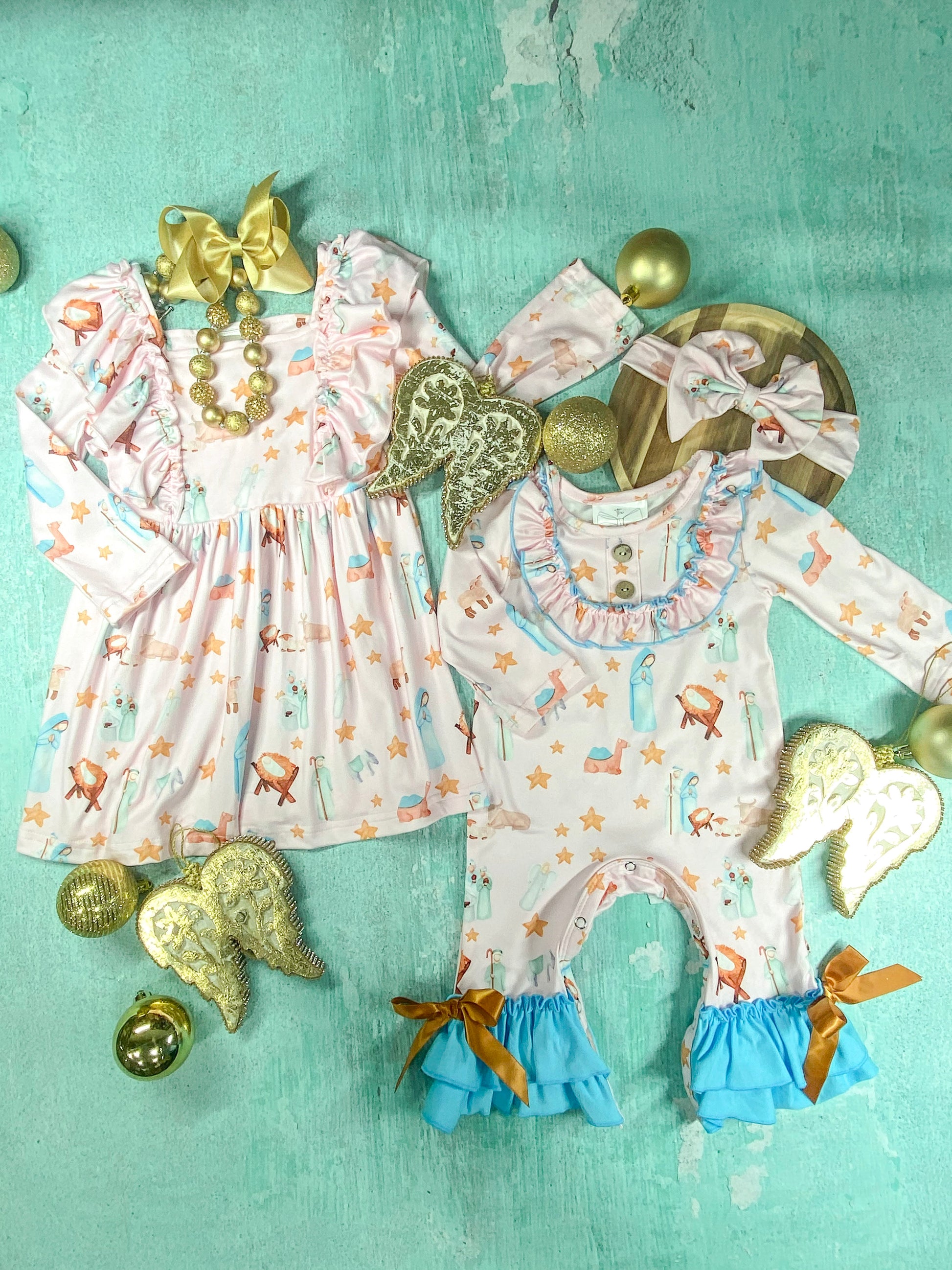 Matching girls’ long sleeve dress and ruffle romper featuring a pastel nativity scene print with gold star accents, displayed with gold Christmas ornaments and accessories on a teal background.