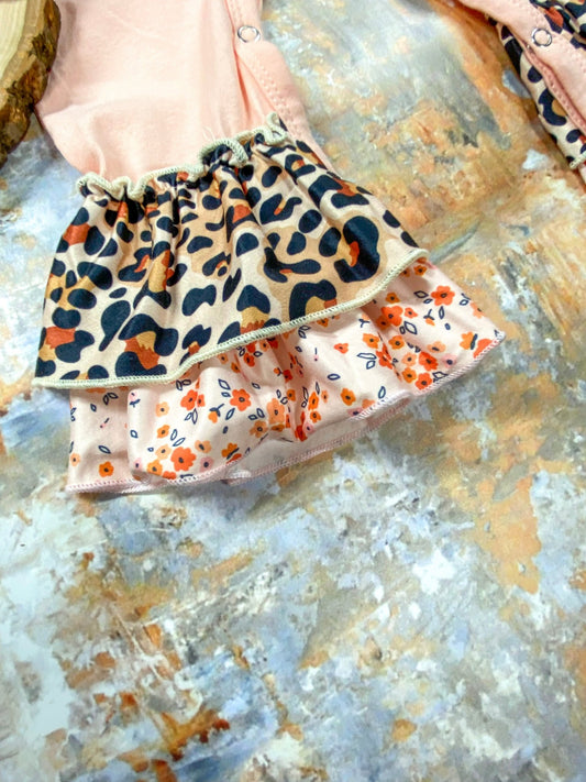 Close-up of the leg cuff on a pink baby romper featuring double-layered ruffles—top layer in a tan leopard print and bottom layer in a peach floral pattern with small orange flowers and blue leaves, displayed on a textured surface. - Alternative view