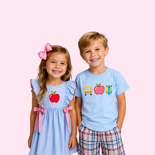 a girl wearing a blue dress with red apple design and pink accents and a boy wearing a blue shirt with school supplies design and plaid shorts on a pink background - Alternative view