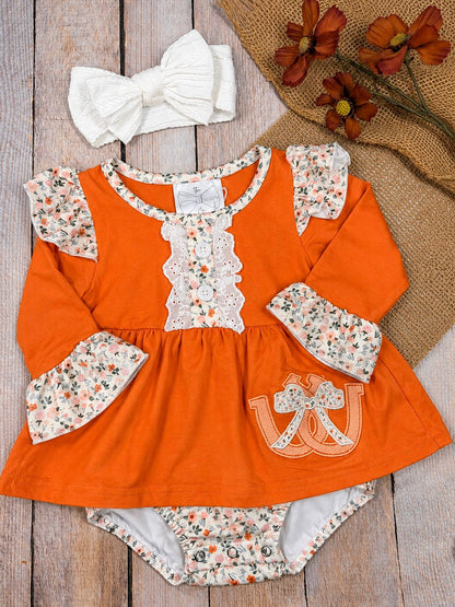 baby girl fall one-piece with horseshoe appliqué, lace ruffle bib, and floral cuffs on a wooden background