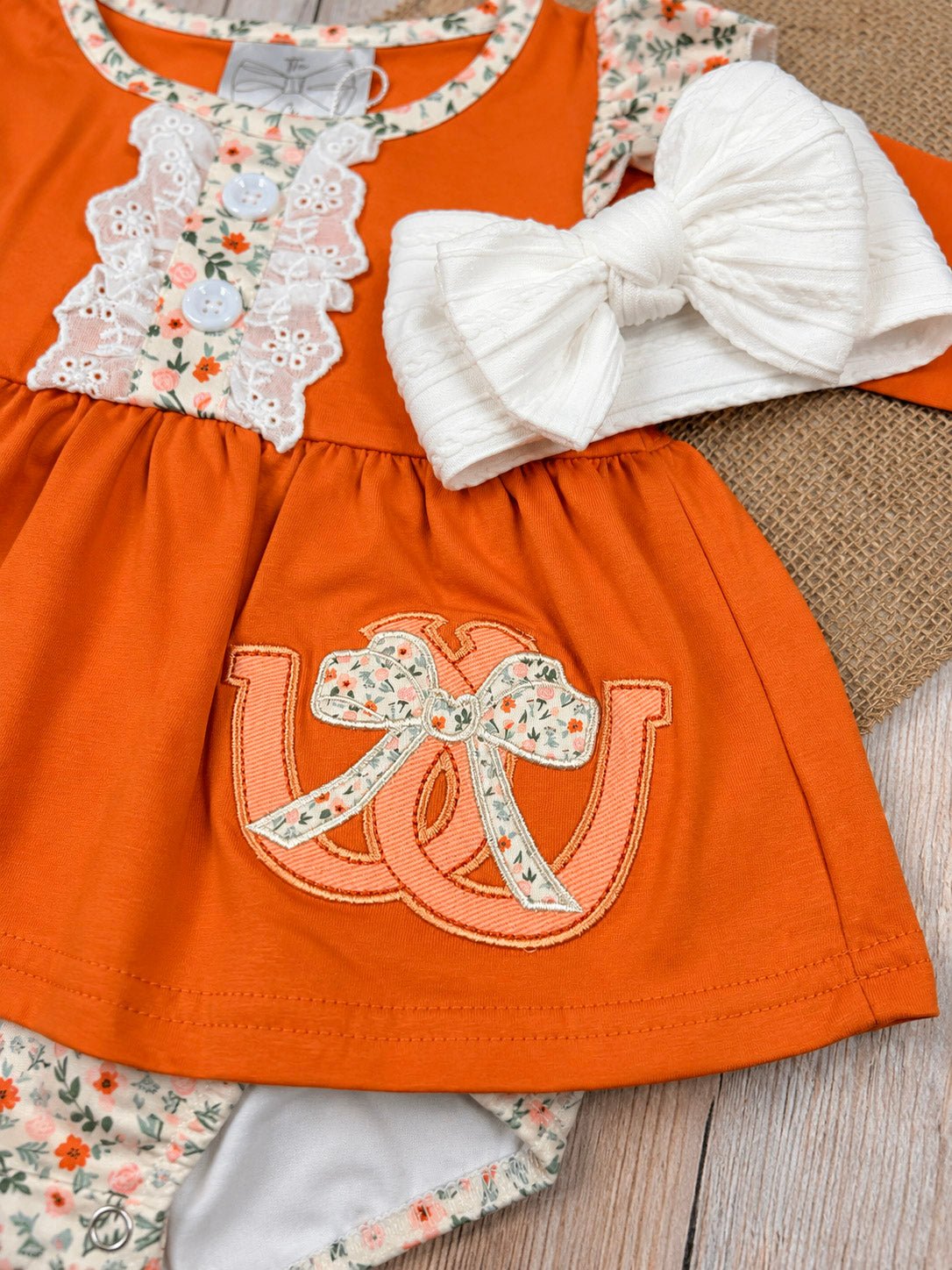 baby girl fall one-piece with horseshoe appliqué, lace ruffle bib, and floral cuffs on a wooden background