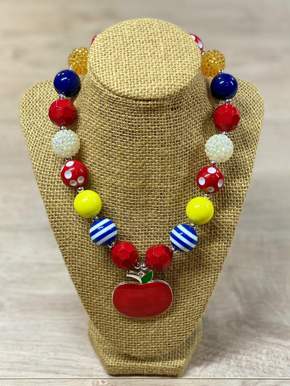 Red apple pendant chunky bead necklace with red polka dot, blue and white striped, white rhinestone, solid red, blue, and yellow beads, with a heart toggle closure.