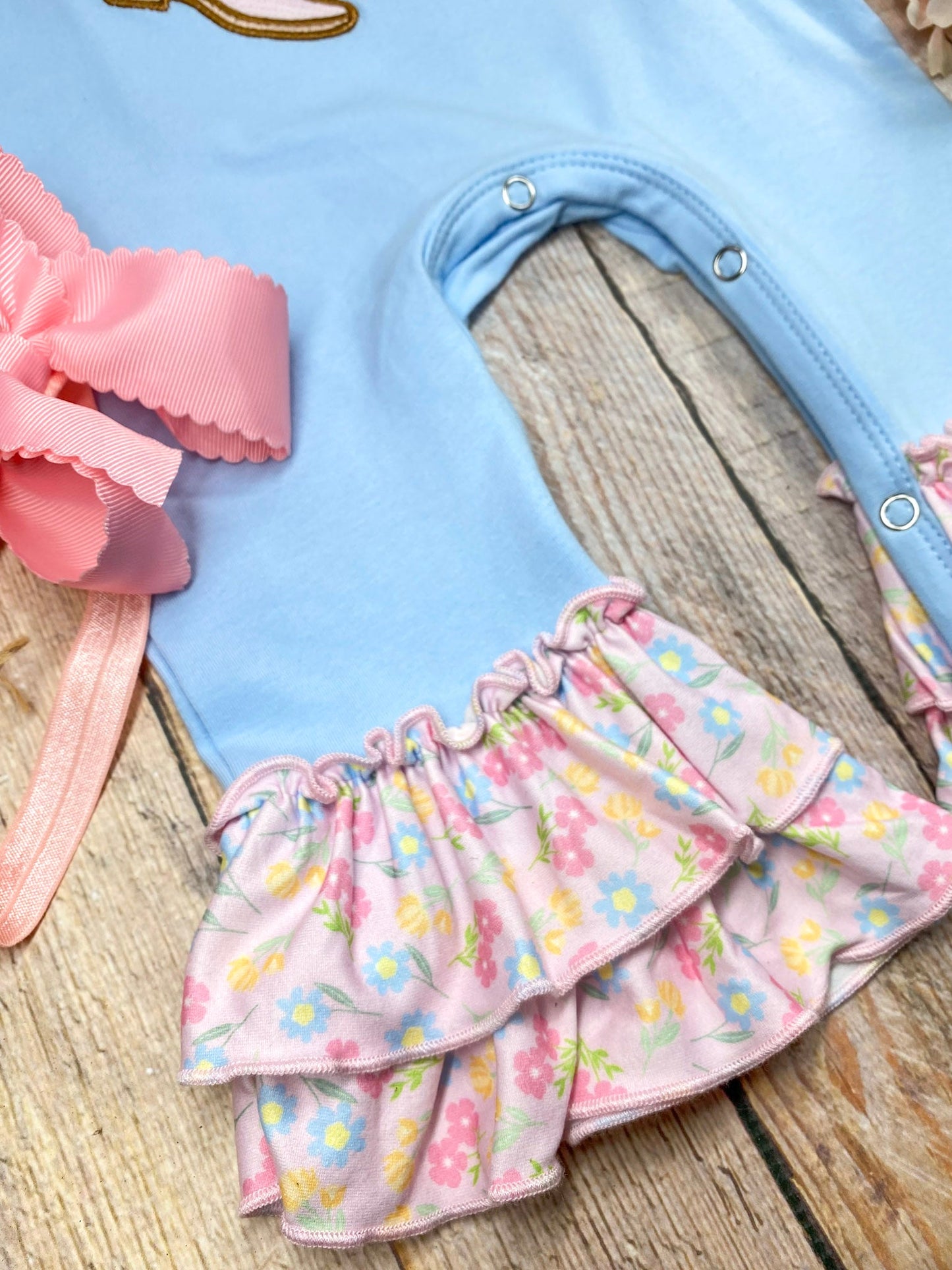 baby girl cowgirl one-piece with boots bow appliqué and floral ruffle leg hems on a wooden background