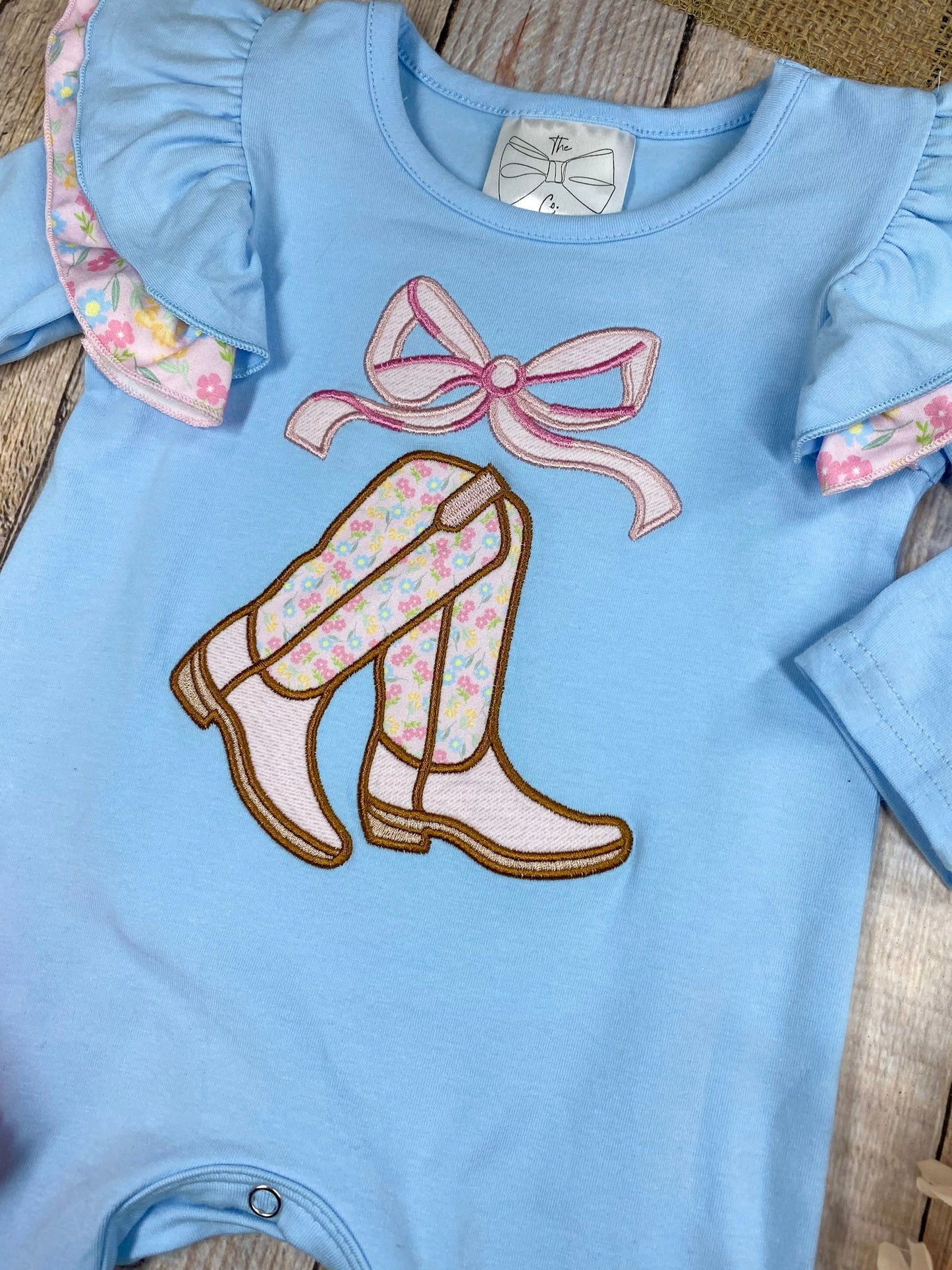baby girl cowgirl one-piece with boots bow appliqué and floral ruffle leg hems on a wooden background
