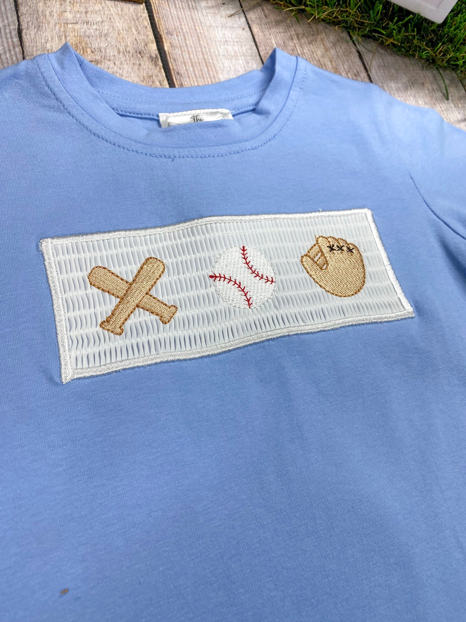 Blue shirt with sports-themed patch featuring a baseball bat, ball, and glove on a wooden surface.
