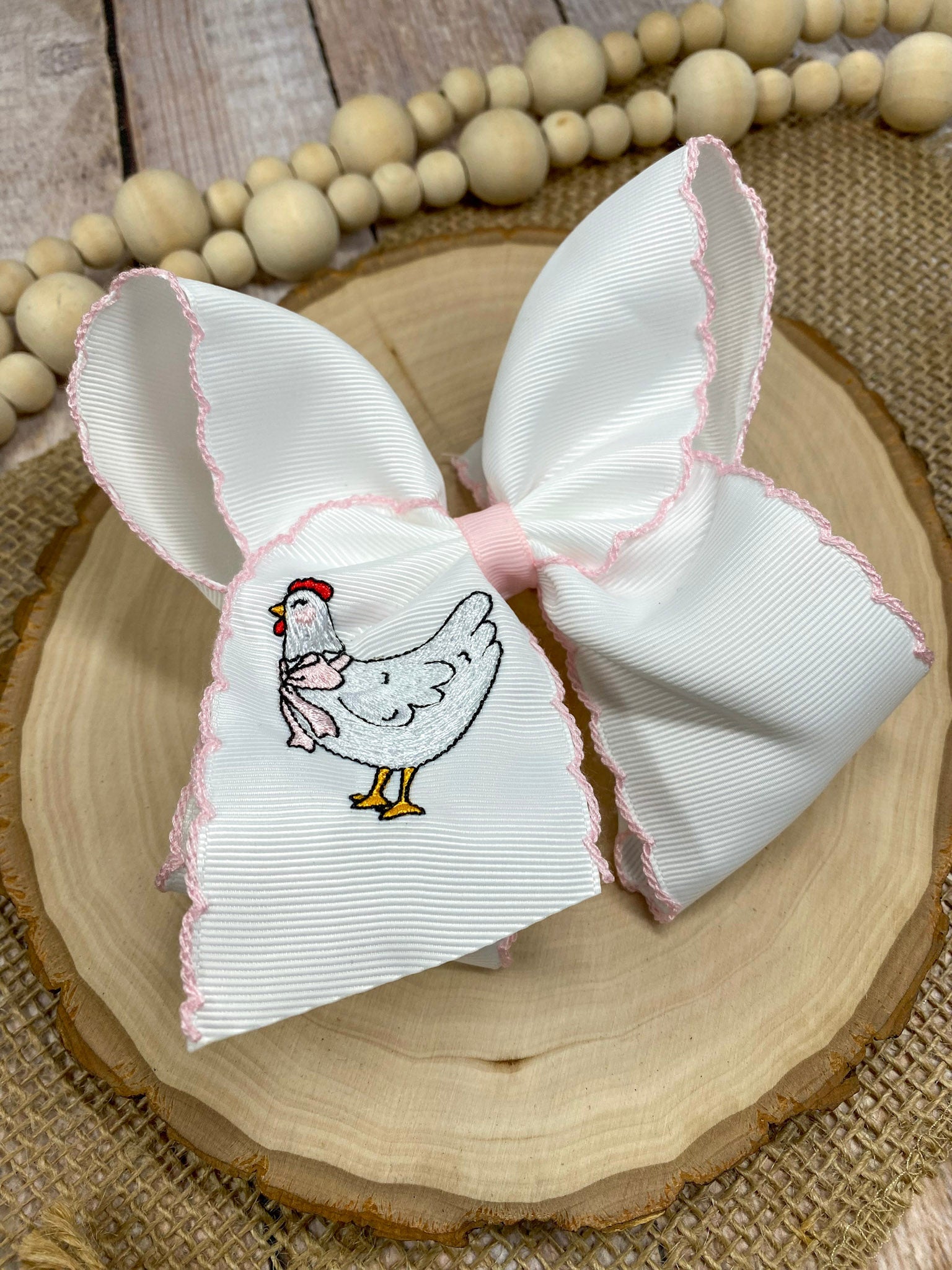 White bow with pink trim featuring an embroidered chicken design on a wooden surface.