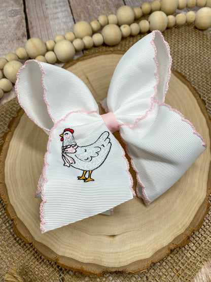White bow with pink trim featuring an embroidered chicken design on a wooden surface.