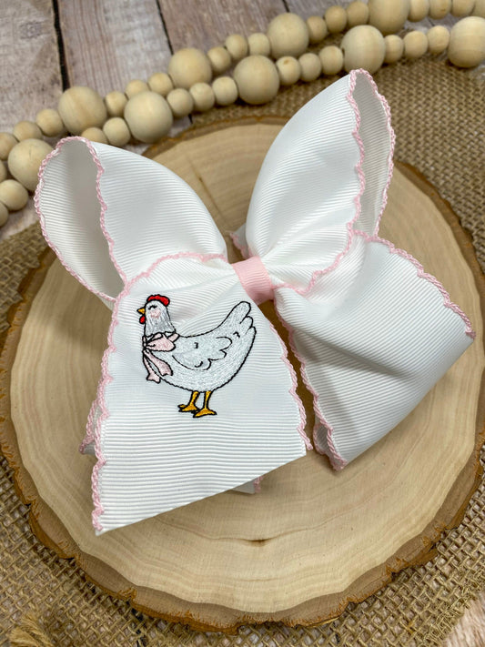 White bow with pink trim featuring an embroidered chicken design on a wooden surface. - Alternative view
