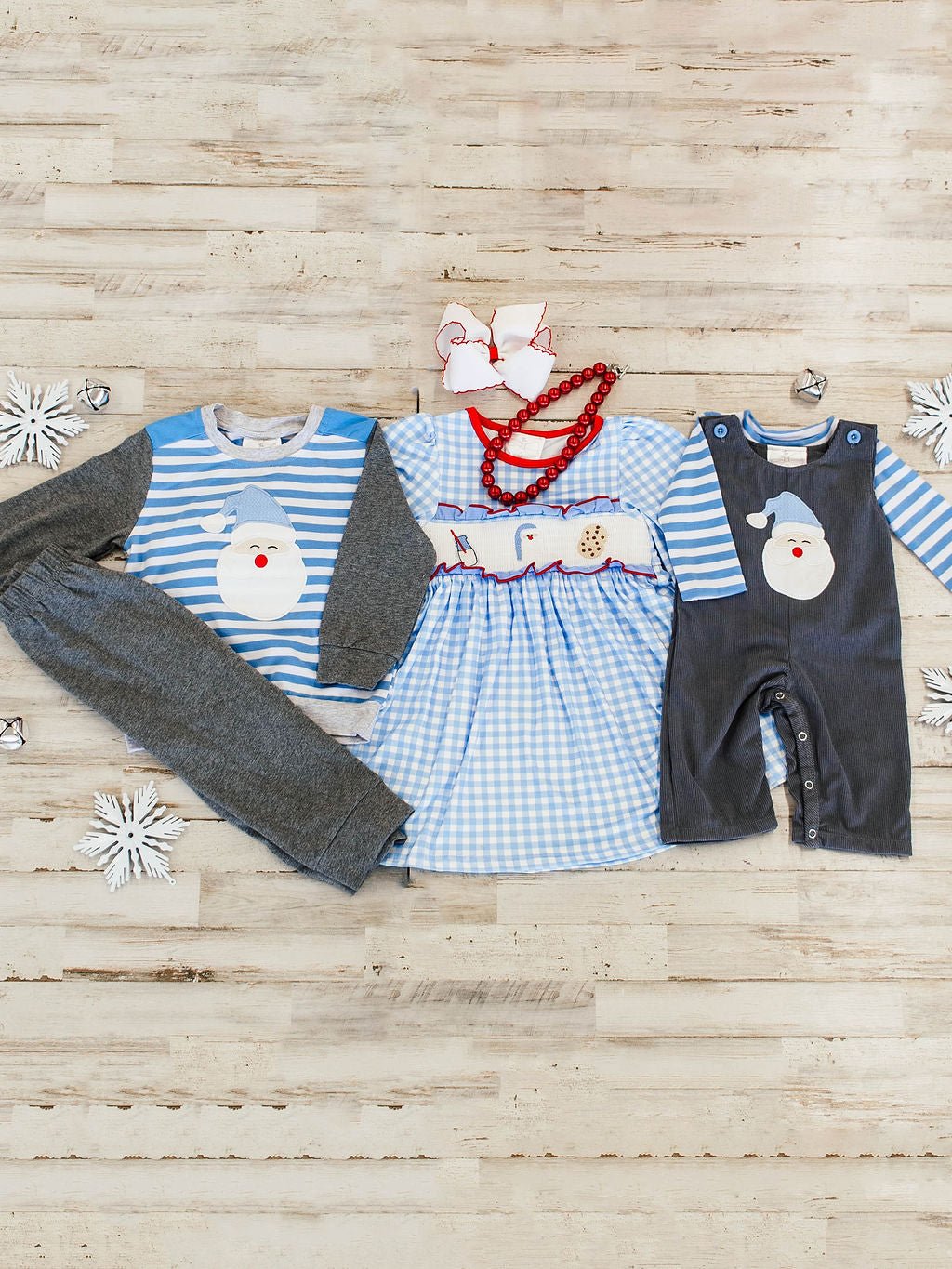 flat lay of sibling sets with far left outfit being a matching boys pants set with striped blue and white long sleeve with a santa wearing light blue hat applique with dark grey sleeves and matching dark grey pants, with girls blue and white gingham dress featuring santa, milk, and cookies embroideries across chest and far right shows the infant overall set with blue and white striped long sleeve with dark grey corduroy overalls on top with a santa applique wearing a light blue hat