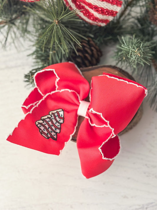 A red embroidered hair bow with a Christmas tree cake design, displayed on a white surface with pine branches and decorative heart-shaped embellishments in the background.