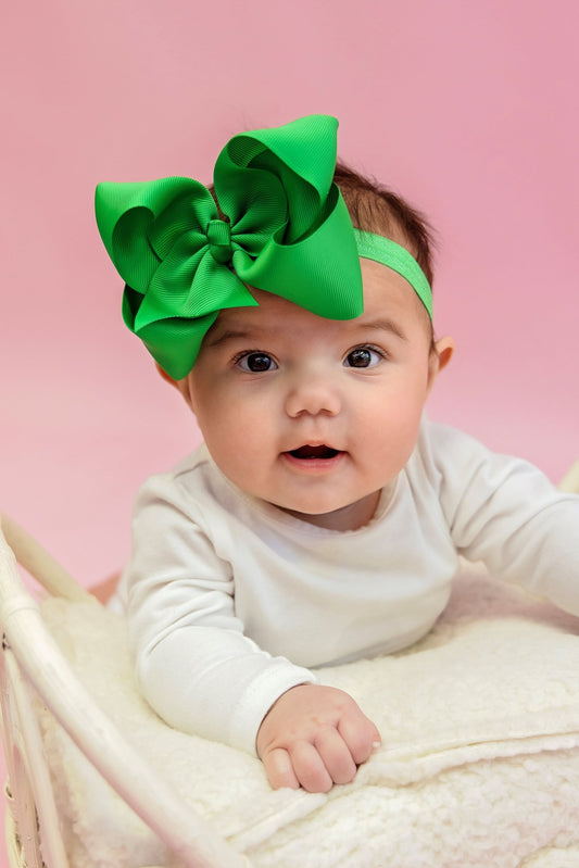 A baby wearing a white outfit and a large green satin hair bow on a pink background.