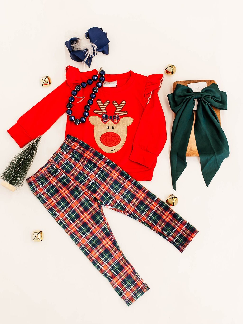 flat lay of girls christmas outfit featuring a red long sleeve with rudolf applique and ruffled shoulders paired with red and green plaid leggings; decorated by a gree coquette bow, navy chunky bead necklace and christmas decorations