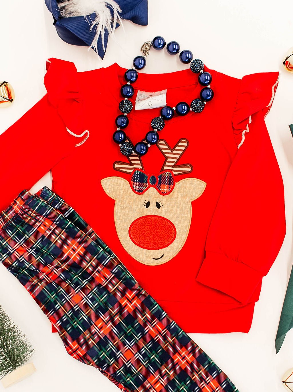 close up of the rudolf applique in center of red long sleeve showing striped antlers and a red and green plaid bow on rudolf that matches the plaid leggings and styled with a navy chunky bead necklace