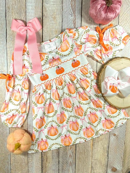 A long-sleeve dress with a white base and a pattern of orange pumpkins tied with pink bows, arranged within green leafy wreaths. The dress features smocking across the chest with embroidered orange pumpkins and pink bows. The sleeves have orange ribbon ties at the elbows. Beside the dress, there is a large pink hair bow with scalloped edges and a smaller white bow with a pumpkin embroidery placed on a wooden round slice (sold separately)