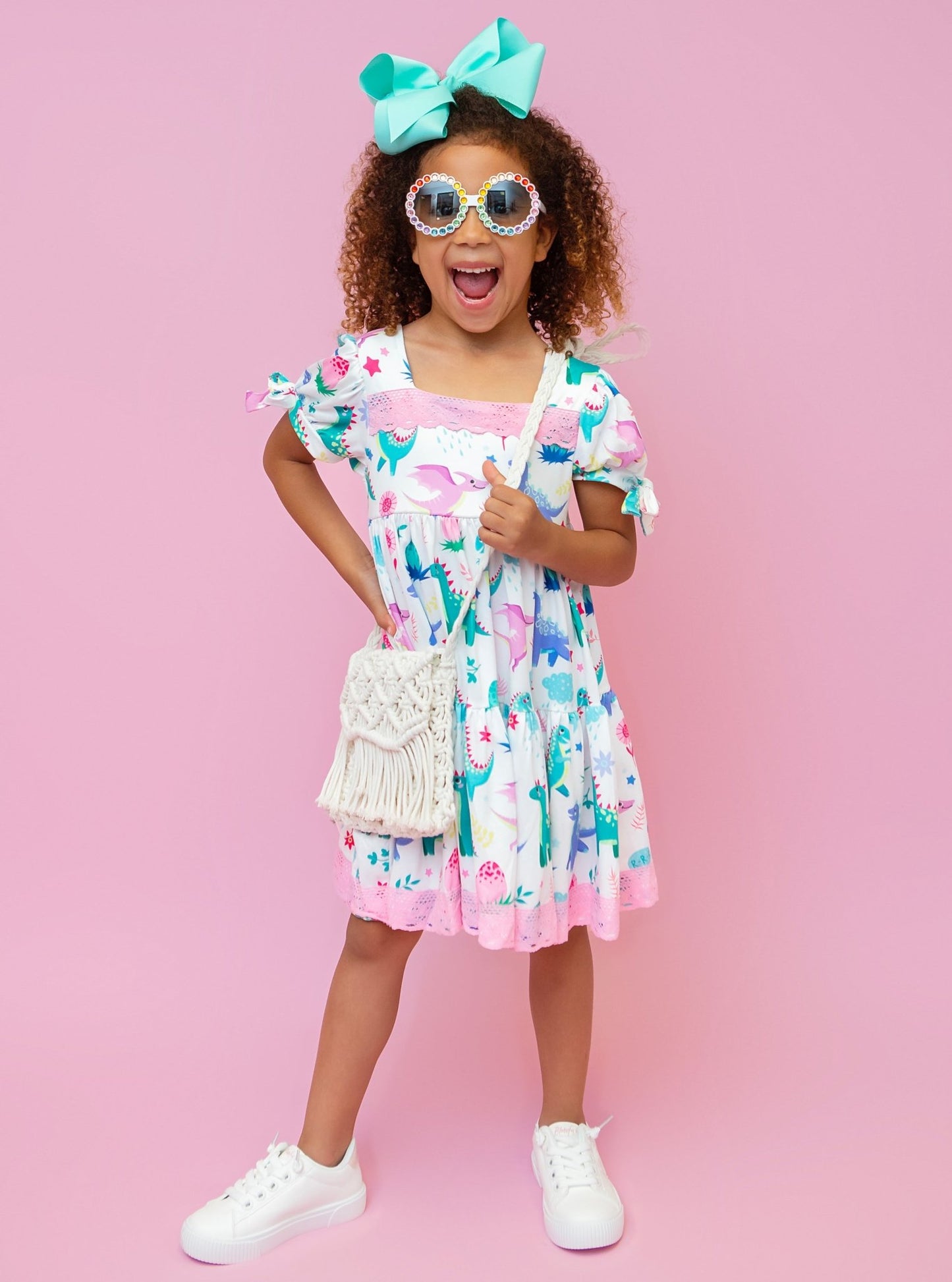 A child model wearing a short-sleeved dress with a dinosaur and floral pattern, accessorized with a bow tie and white sneakers.