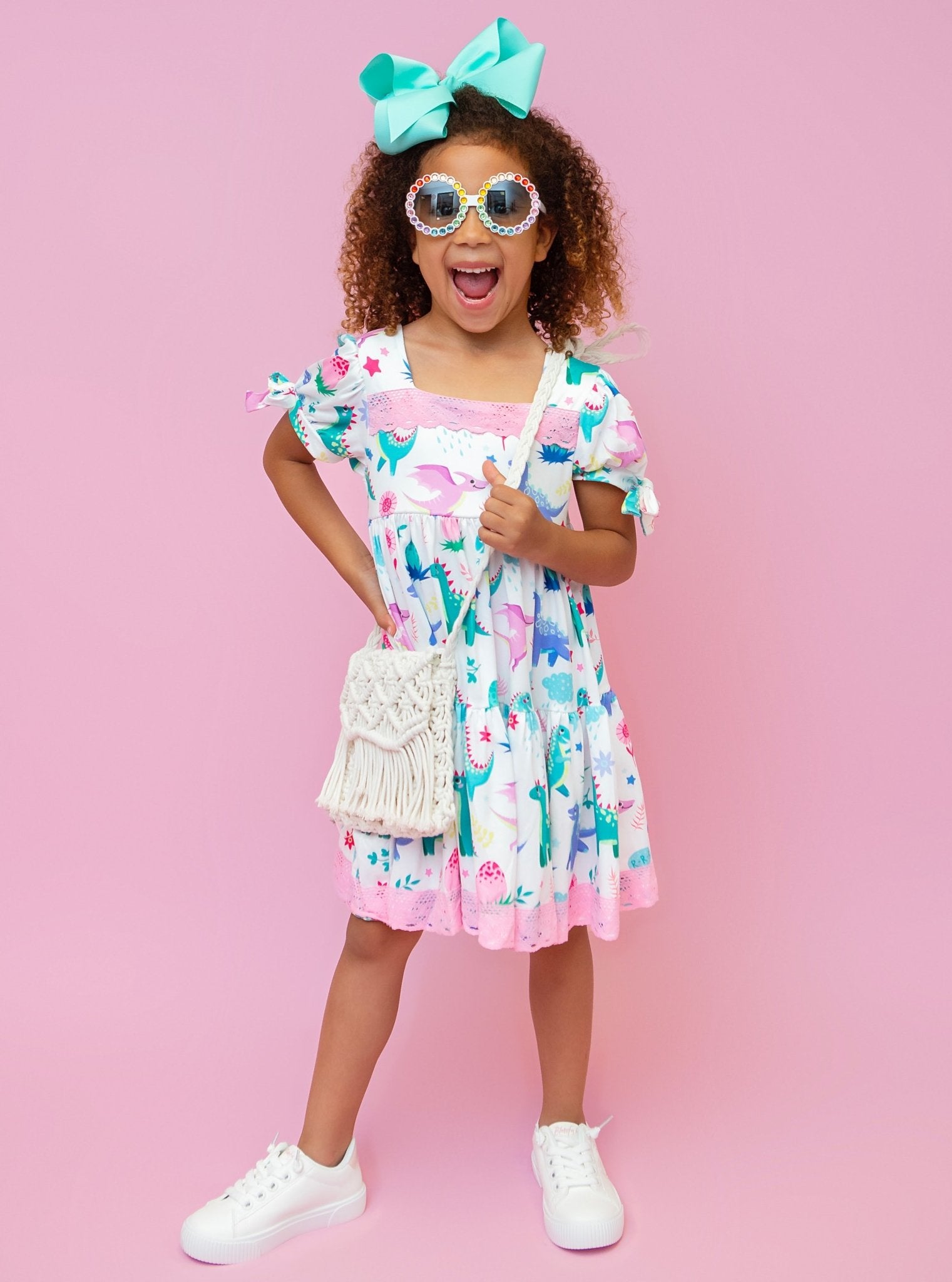 A child model wearing a short-sleeved dress with a dinosaur and floral pattern, accessorized with a bow tie and white sneakers.