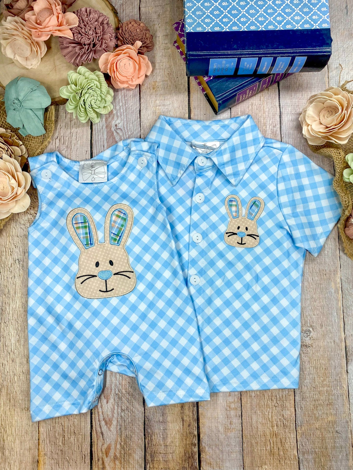 Blue checkered baby romper with bunny design on a wooden surface with flowers.