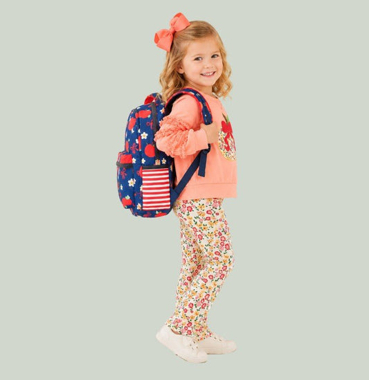 A young girl with blonde hair and a coral bow headband is shown from the side, wearing a peach sweatshirt, floral leggings, and white sneakers, while carrying a navy blue backpack decorated with a red and white American flag and white flowers. The background is a solid light sage green. - Alternative view