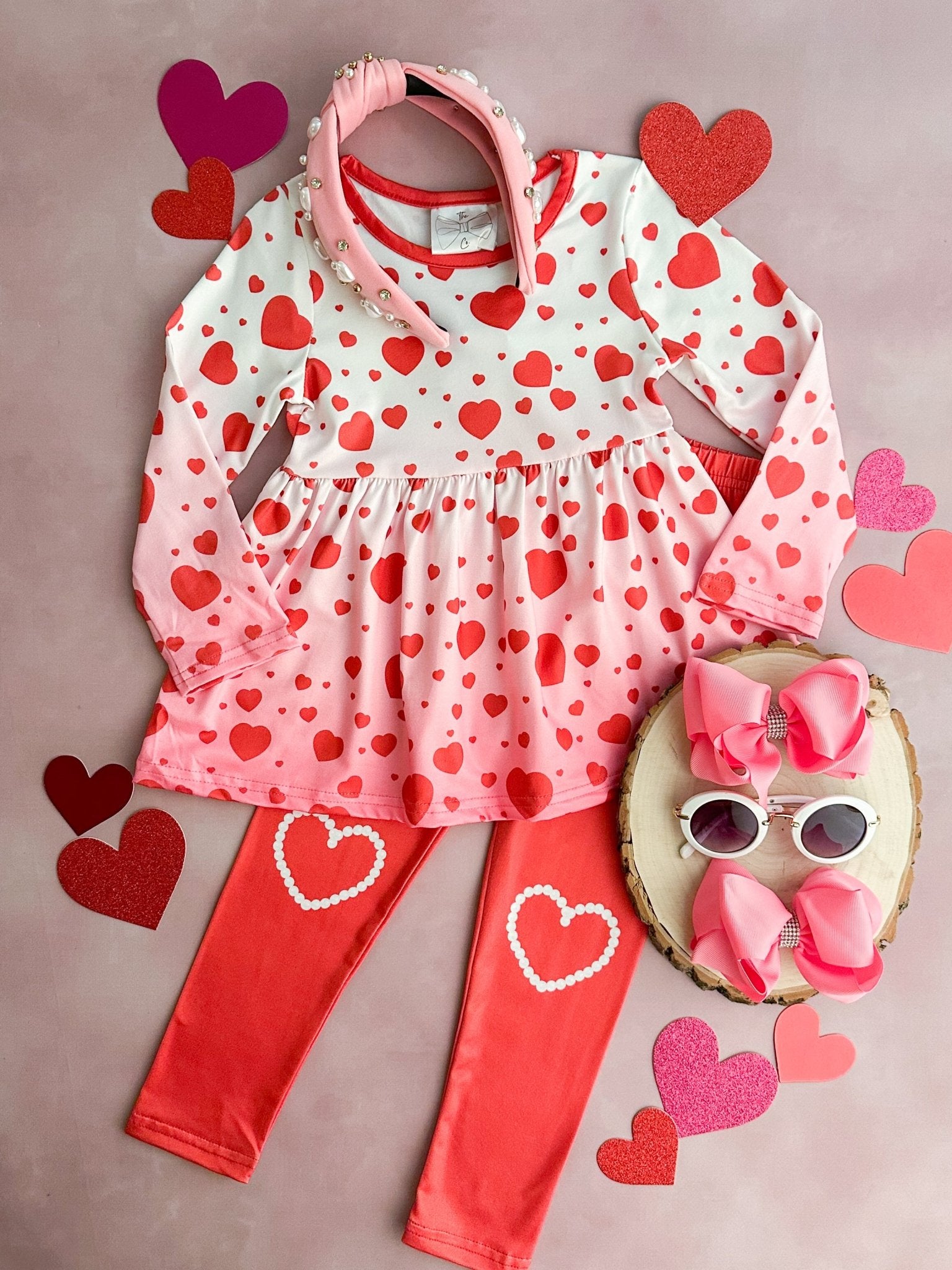 A pink and white ombre leggings set for toddlers, with red hearts and a bow, displayed alongside pink sunglasses and a heart-decorated wooden clipboard.