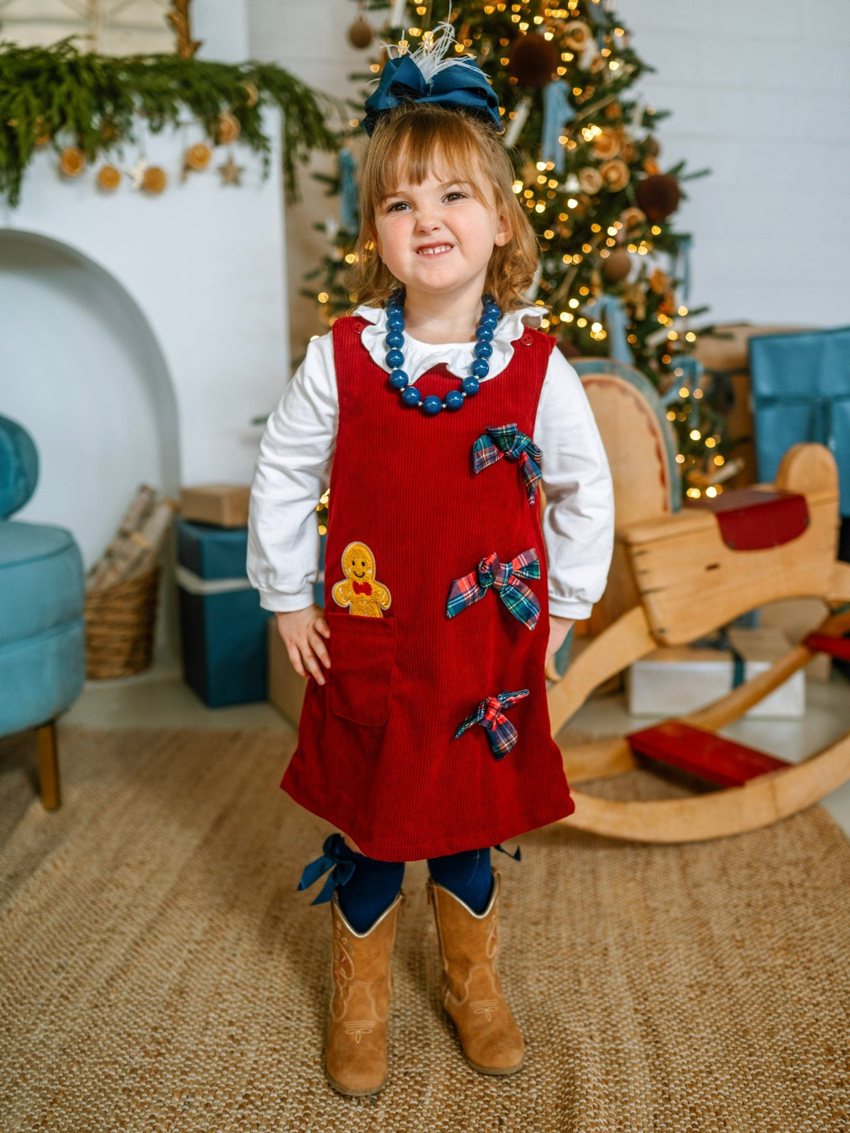 Girl wearing a red gingerbread corduroy jumper dress by the Christmas tree