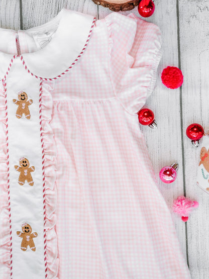Pink baby dress with gingerbread man embroidery on a wooden surface with Christmas decorations.