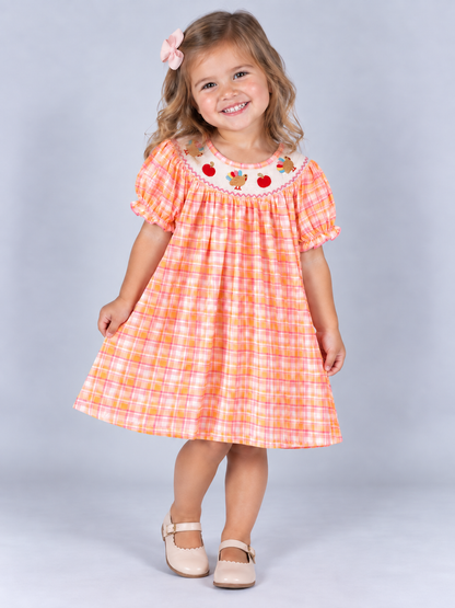 model wearing Pink and orange plaid girls’ Thanksgiving dress with a smocked neckline, embroidered turkeys and pumpkins, and ruffled sleeves