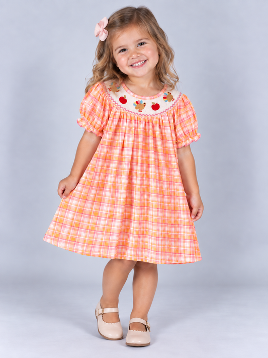 model wearing Pink and orange plaid girls’ Thanksgiving dress with a smocked neckline, embroidered turkeys and pumpkins, and ruffled sleeves
