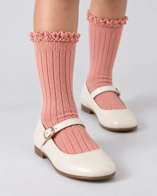 A pair of pink ribbed knee-high socks with ruffled trim, worn with cream Mary Jane shoes on a light gray studio background.
