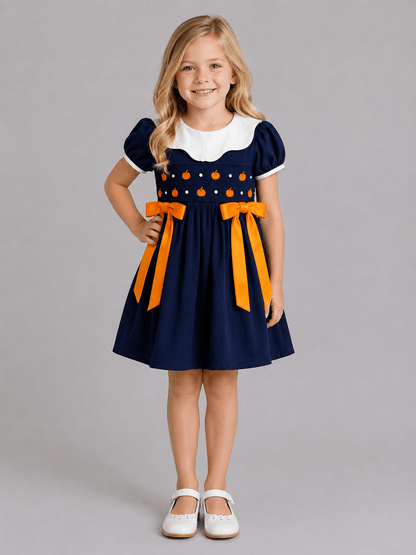 Navy girls fall dress with pumpkin smocking, white Peter Pan collar and orange bows on a rustic autumn background.