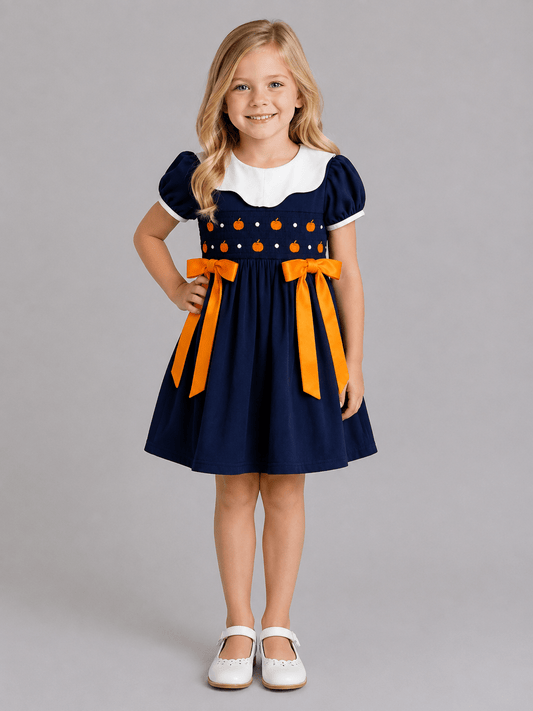 Navy girls fall dress with pumpkin smocking, white Peter Pan collar and orange bows on a rustic autumn background.
