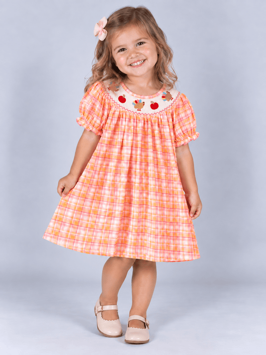model wearing Pink and orange plaid girls’ Thanksgiving dress with a smocked neckline, embroidered turkeys and pumpkins, and ruffled sleeves