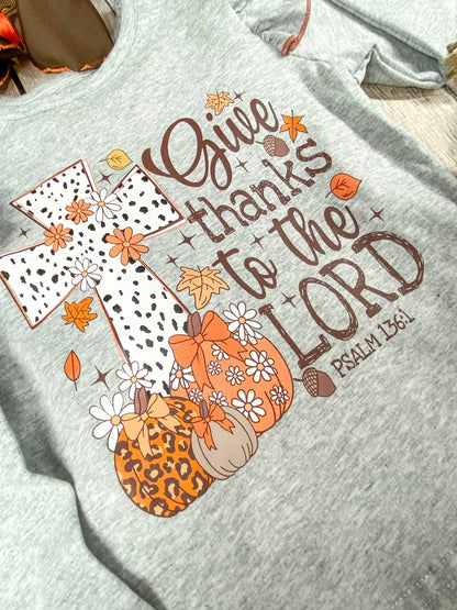 close up of the print text saying "Give thanks to the Lord Psalm 136:1" with a cross with some pumpkins in front of the cross on grey long sleeve