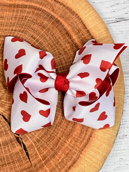 Valentine heart print white hair bow with red bow on wooden background – cute girls accessory from The Hair Bow Company