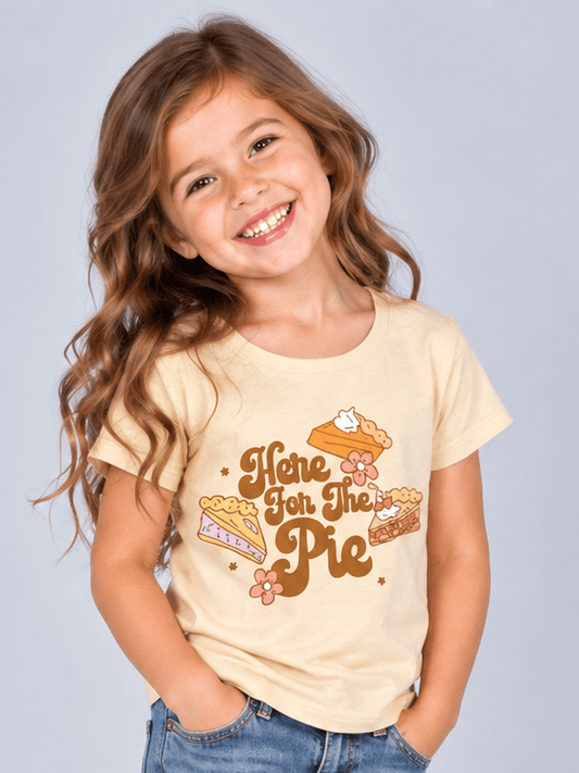 A young girl with long brown hair stands smiling in a studio, wearing a light beige graphic T-shirt that reads 'Here for the Pie' with a pie graphic, blue jeans, and brown cowboy boots against a light gray background.