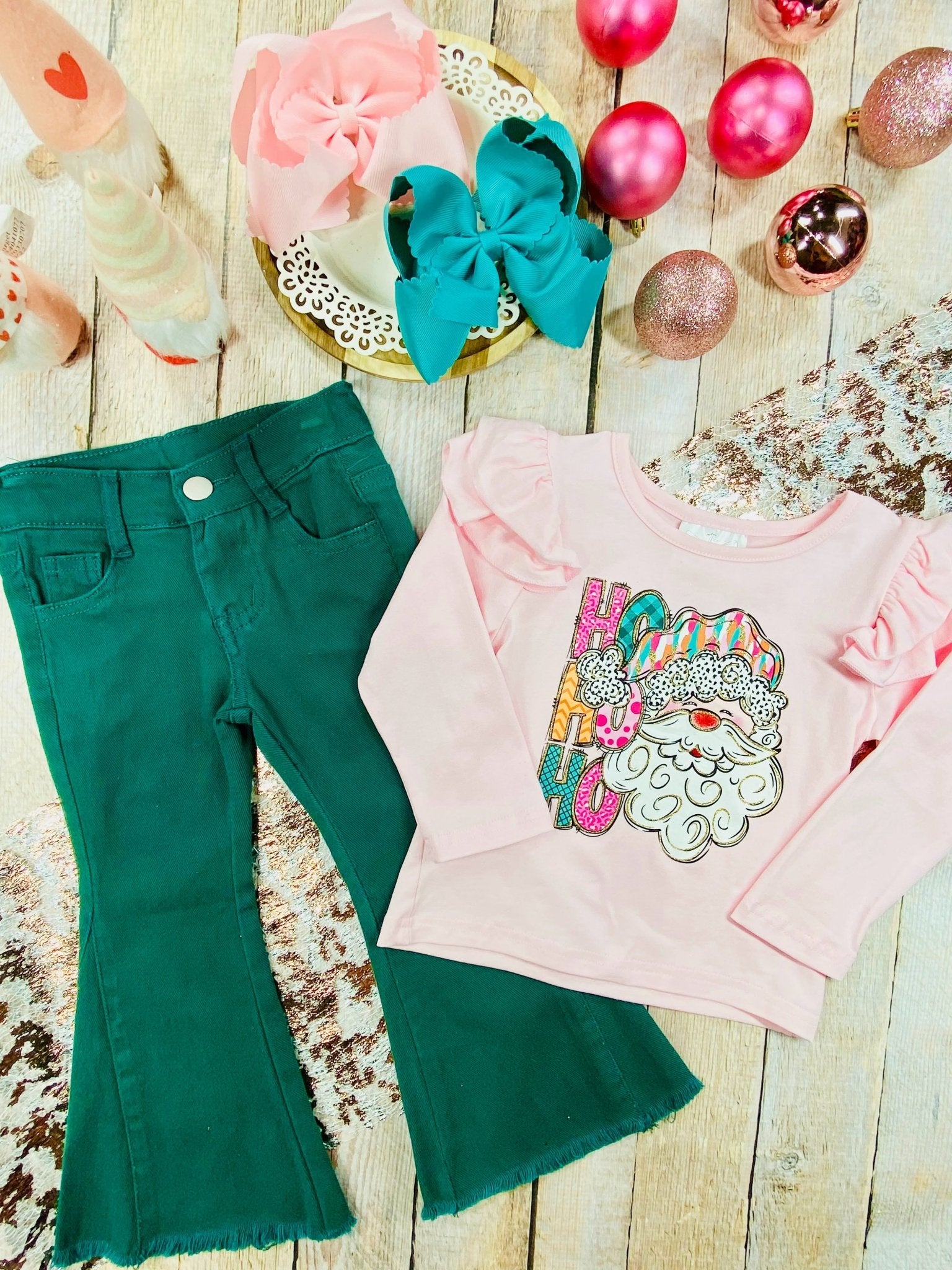 Toddler girl holiday outfit consisting of a light pink long-sleeve top with ruffled shoulders and a colorful Santa graphic that says “Ho Ho Ho,” paired with dark green bell-bottom pants featuring raw, frayed hems. The shirt has a playful, festive design with bright colors and glitter details, and the outfit has a cute, coordinated Christmas look