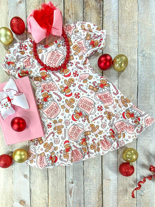 Full flat lay of a girls’ Christmas dress with puff sleeves and a playful holiday print of gingerbread cookies, candy canes, hot cocoa mugs reading “Merry & Bright,” and snowflakes on a cream background, styled with a pink bow, red bead necklace, gift box, and festive ornaments.