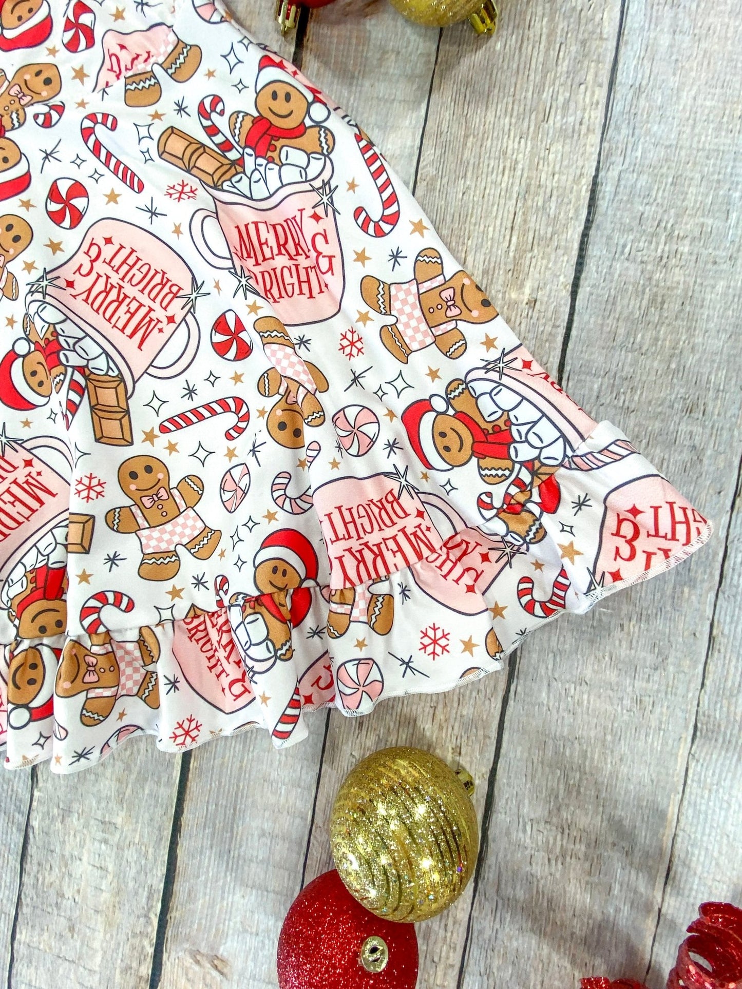 Detailed view of the ruffled hem of a holiday girls’ dress decorated with cheerful gingerbread figures, candy canes, snowflakes, and “Merry & Bright” cocoa mugs, styled with glittery red and gold Christmas ornaments on a wood background.