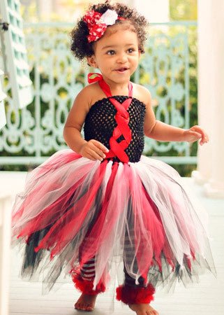 A young toddler girl wearing a colorful costume with a black crochet top featuring red ribbon crisscrossing in the front. Her skirt is made of layered tulle in red, black, and white, giving it a tutu-style. She also has matching accessories, including a red and white flower headband and striped leggings. She is smiling and standing on a porch with decorative railings in the background
