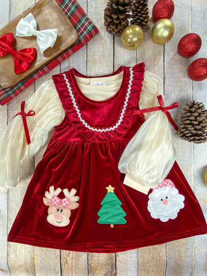 Flat lay of a girls’ red velvet Christmas dress with cream puff sleeves and red ribbon ties at the wrists. The dress features festive appliqués of a reindeer with a plaid bow, a green Christmas tree, and Santa Claus with a red checkered hat. It has a ruffled neckline with white trim and is displayed with pinecones, ornaments, and red and white hair bows on a plaid tray