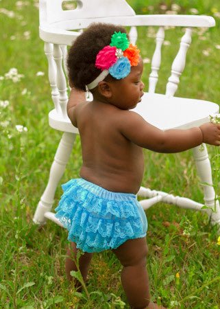 A baby wearing a blue lace diaper cover and a headband with multicolored flowers is standing by a white chair outdoors.