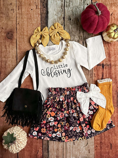 Children's outfit with floral skirt, white top, and accessories on a wooden surface.
