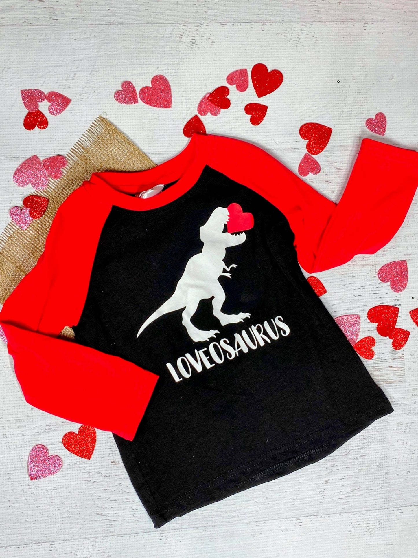 A long sleeve black t-shirt with a white dinosaur graphic and the text 'LOVEOSAURUS' on the front, displayed on a wooden surface with red heart decorations around it.