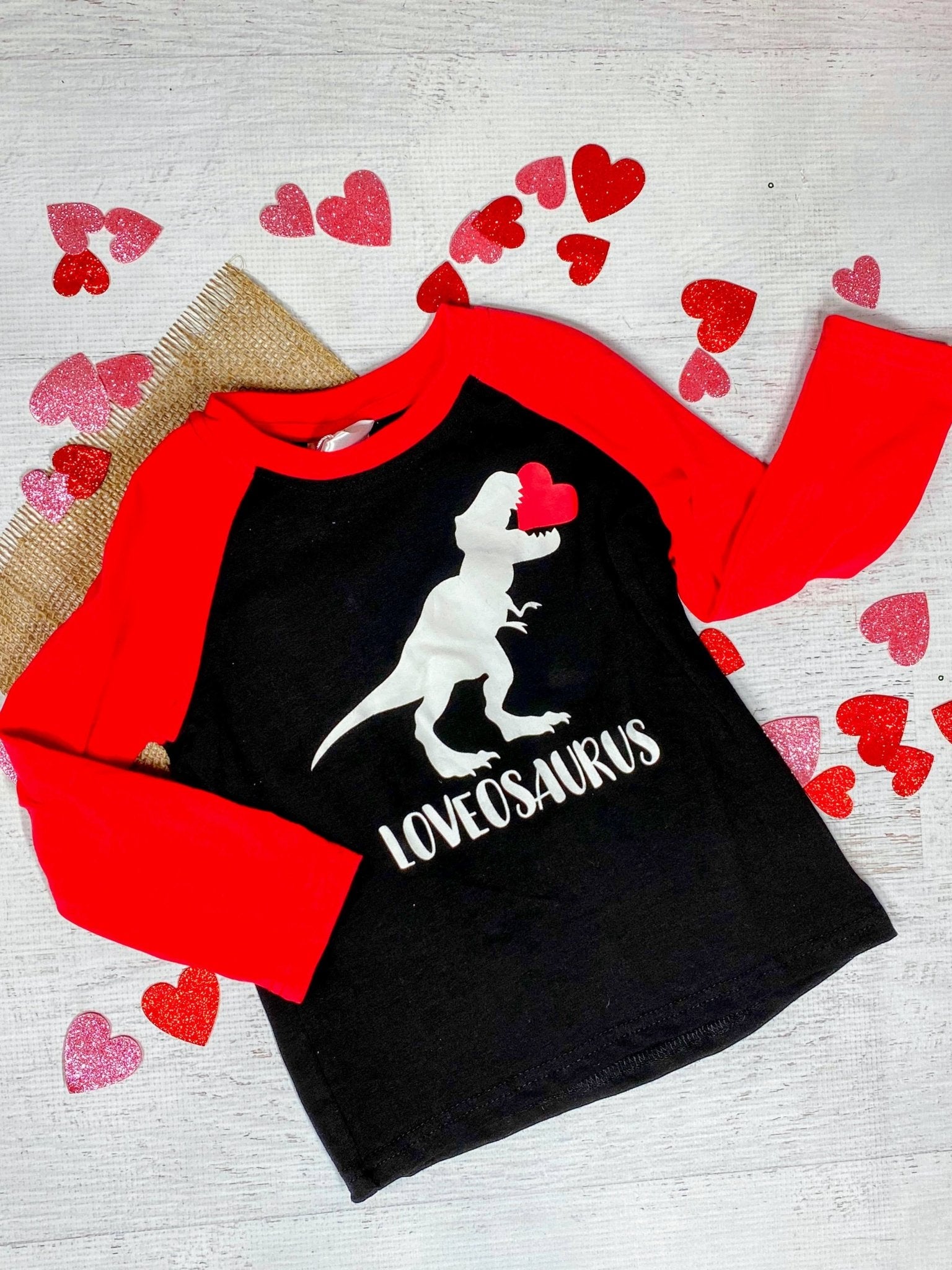 A long sleeve black t-shirt with a white dinosaur graphic and the text 'LOVEOSAURUS' on the front, displayed on a wooden surface with red heart decorations around it.