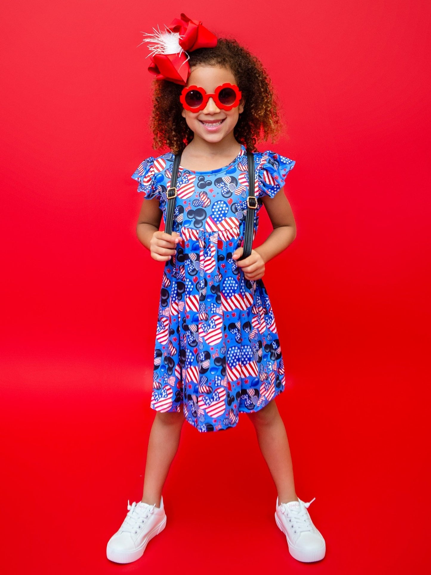 A girl wearing a blue dress with a Mickey and Minnie Mouse patriotic design, featuring an American flag motif, and red accessories, standing against a red background.