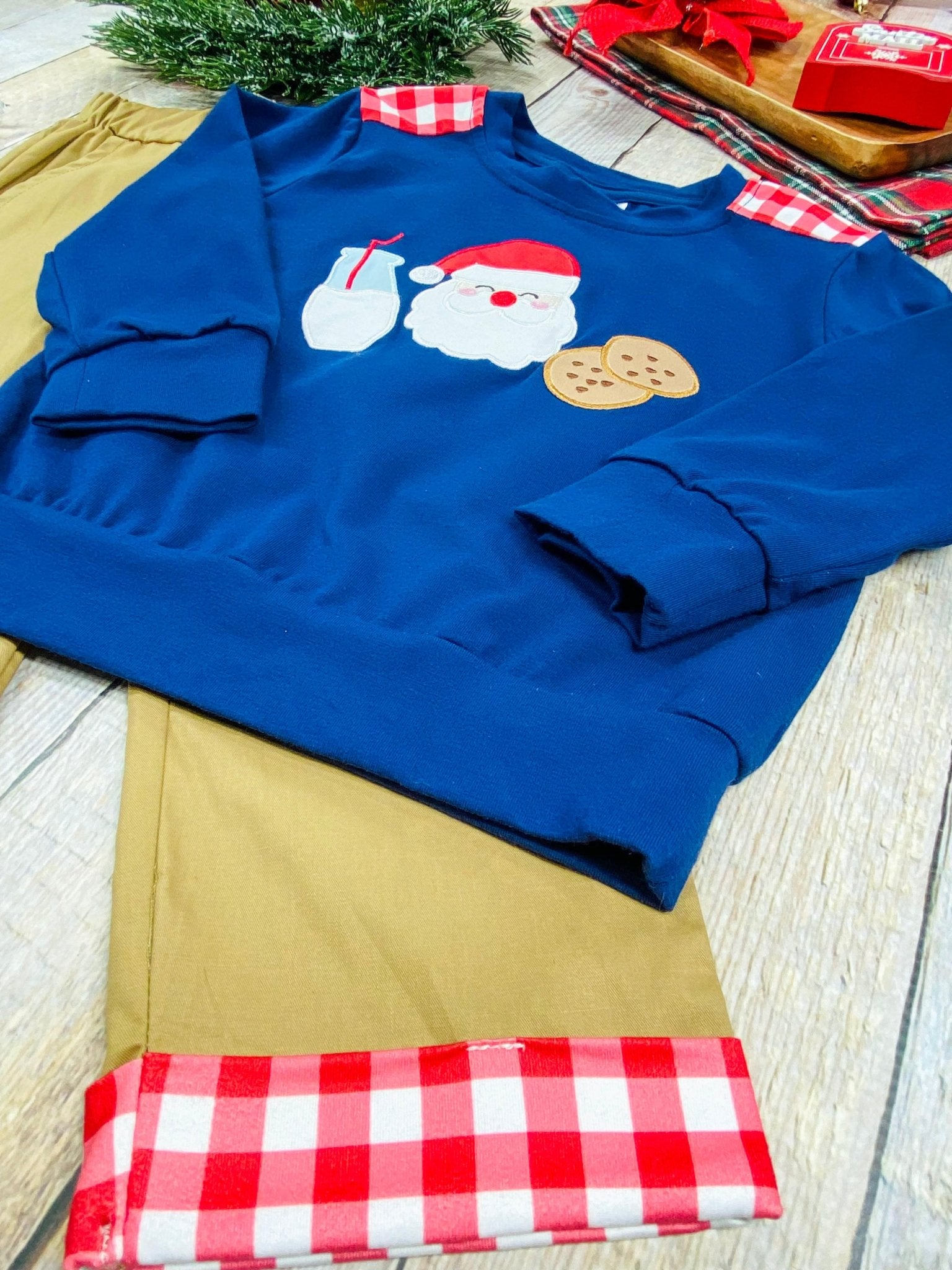 Close-up of a boys’ Christmas outfit featuring a navy blue sweatshirt with Santa, milk, and cookies applique design, paired with tan pants trimmed in red and white buffalo plaid