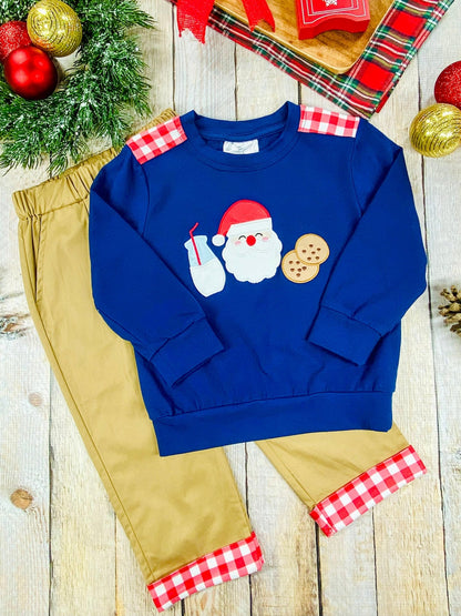 Flat lay of a boys’ holiday outfit with a navy blue sweatshirt decorated with Santa, milk, and cookies applique, styled with tan pants that have red and white plaid cuffs