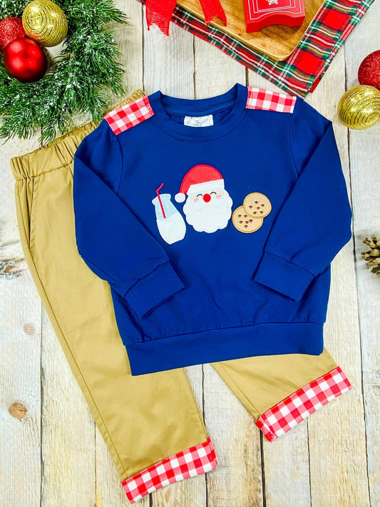 Flat lay of a boys’ holiday outfit with a navy blue sweatshirt decorated with Santa, milk, and cookies applique, styled with tan pants that have red and white plaid cuffs