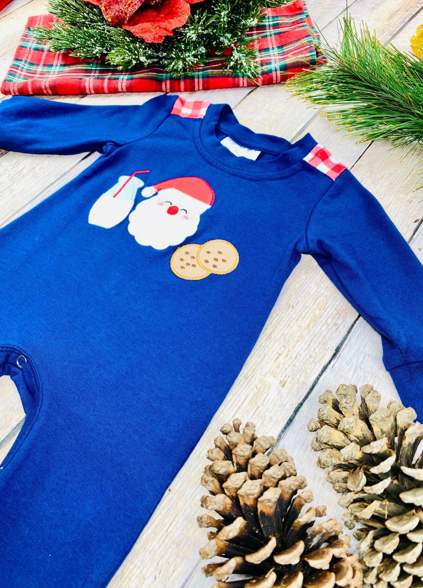A navy blue baby romper photographed at an angle on a light wooden surface, featuring red-and-white plaid accents on the shoulders and a holiday appliqué of Santa’s face, a glass of milk, and two cookies on the chest. The bottom portion of the romper is partly visible, showing some of the metal snaps along the inseam