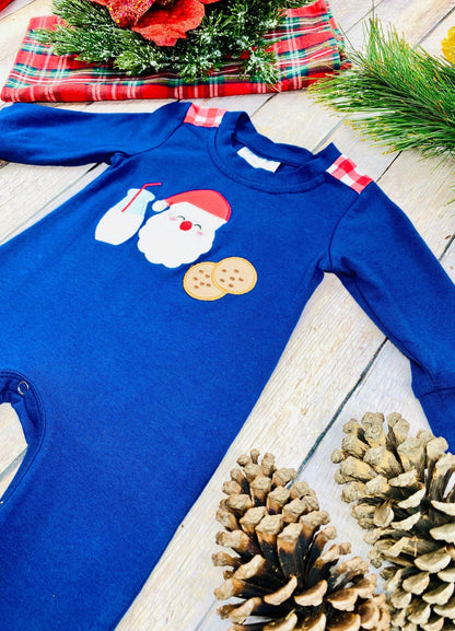 A navy blue baby romper photographed at an angle on a light wooden surface, featuring red-and-white plaid accents on the shoulders and a holiday appliqué of Santa’s face, a glass of milk, and two cookies on the chest. The bottom portion of the romper is partly visible, showing some of the metal snaps along the inseam